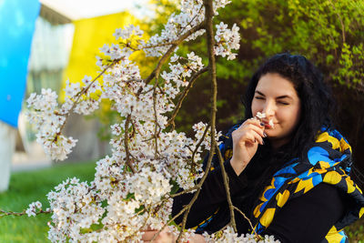Young ukrainian brunette woman enjoying aroma of cherry blossoms while walking in park in spring