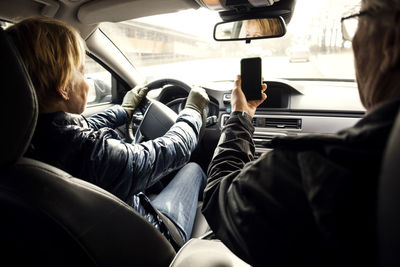 Senior woman driving car while sitting by man taking selfie through smart phone
