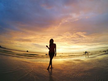 Silhouette woman standing on wet shore at beach against cloudy sky during sunset