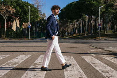 Full body side view of elegant hispanic male walking on pedestrian crossing on sunny street with green trees in city