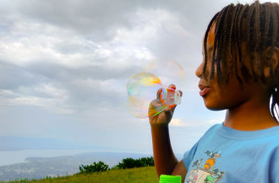 Side view of boy blowing bubbles against cloudy sky