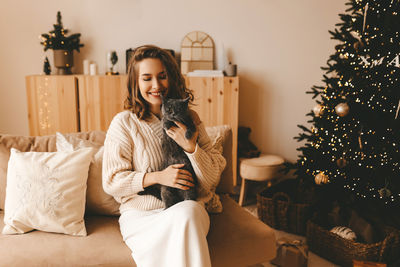Smiling happy girl hugging her pet cat while sitting by a decorated christmas tree in a cozy home