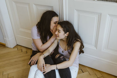 Young woman whispering to female friend against white door at home