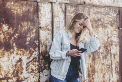 Young woman using mobile phone while standing against rusty wall