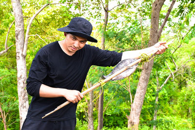 Portrait of young man holding stick with dead fish at forest