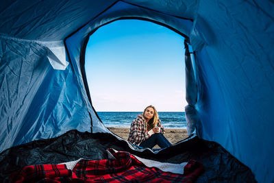 Portrait of woman sitting by tent at beach against sky