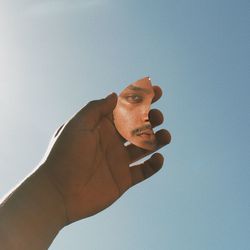 Cropped hand of man holding mirror with his reflection against clear sky