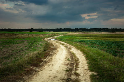 Sandy road through green meadows and fields, dark rainy evening clouds