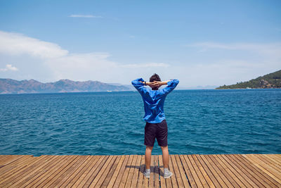 Guy stands rear view by the sea on the pier in a blue shirt in sneakers