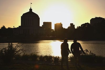 Silhouette men in river by city against sky during sunset