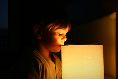 Close-up of boy by illuminated lamp in dark at home