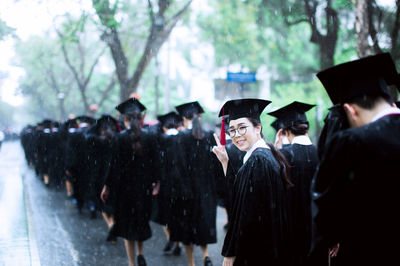 Portrait of happy young woman wearing mortarboard while gesturing peace sign with friends on road during rain