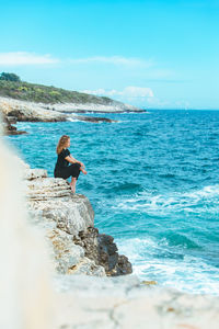 Rear view of woman sitting on rock by sea against sky