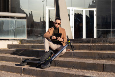 Young handsome bussinesman sitting on stairs in financial district with electric scooter to commute 