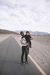 Playful father lifting bicycle with son while standing on country road against mountains and cloudy sky