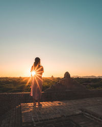 Rear view of young woman looking at landscape while standing on old ruin against clear sky during sunset