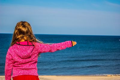 Rear view of girl standing with outstretched arm at beach against sky
