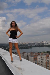 Stylish young woman standing on retaining wall of building terrace against cloudy sky