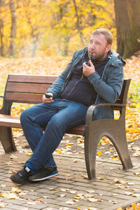Man smoking electronic cigarette while sitting on bench against tree