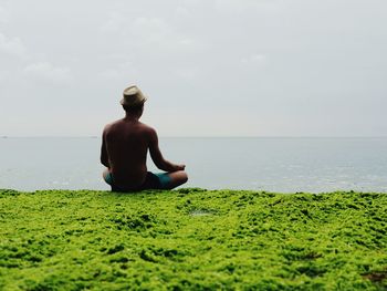 Rear view of man meditating while sitting by sea against sky