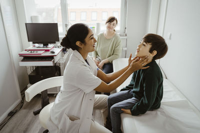 Side view of smiling female pediatrician examining boy sitting on bed in examination room at clinic