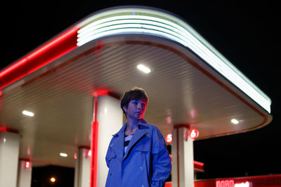 Lonely woman standing against petrol station in red neon light at night, waiting for car to refuel