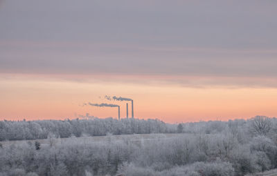 Snow covered trees with smoke stacks in background against cloudy sky during sunset