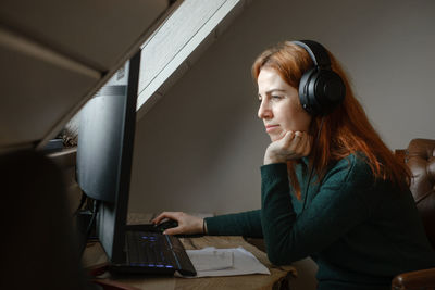 Middle age woman listening to music in big headphones near computer at home. creating play list