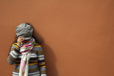Woman covering face with cap while standing by brown wall