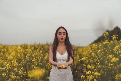 Portrait of young woman standing amidst yellow flowering plants on field against sky