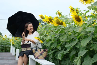 Smiling woman with umbrella while sitting at sunflower field