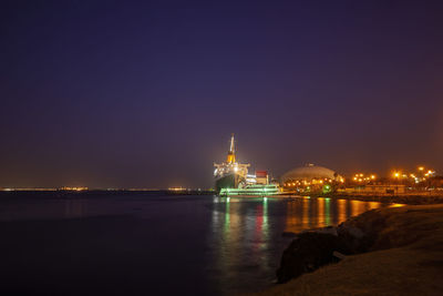 Illuminated buildings by sea against sky at night