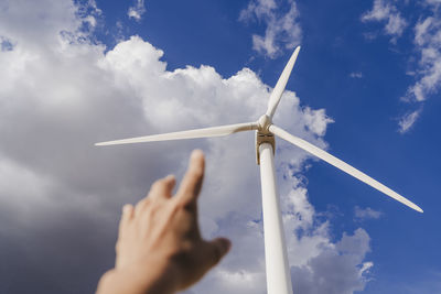 Hand of woman gesturing at rotor of wind turbine below clouds in sky