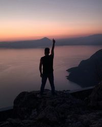 Silhouette man standing with arms raised on cliff by sea at sunset