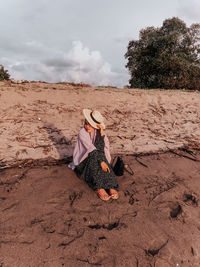 Woman wearing hat sitting at beach against sky