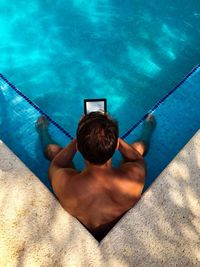 High angle view of shirtless man holding digital tablet while sitting in swimming pool