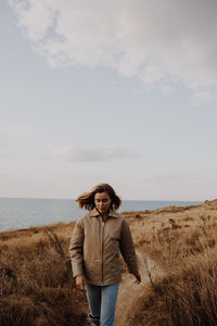 Young woman walking on dirt road against sea and sky