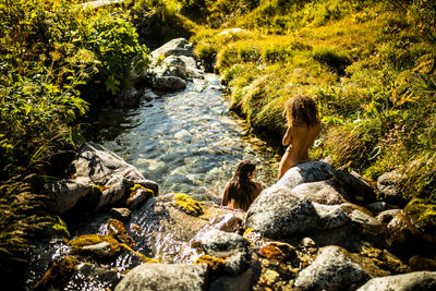 From above back view of unrecognizable female travelers sitting in water of narrow stony stream flowing among green bushes in summer day in pyrenees mountains