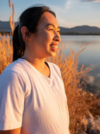 Side view of happy hopeful woman looking away to nature reflection on water at sunset lakeside.