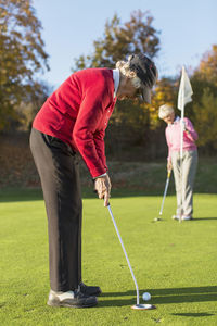 Side view of senior woman putting while friend holding flag on golf course