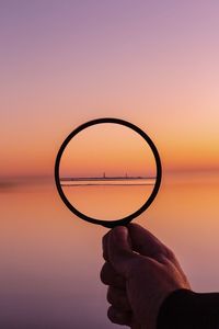 Silhouette hand holding magnifying glass at sea against sky during sunset