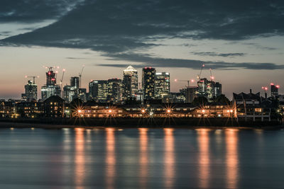 Illuminated buildings by river against sky in city