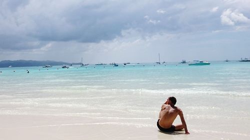 Rear view of man sitting on shore at beach against cloudy sky