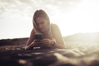 Young woman using smart phone at beach against sky