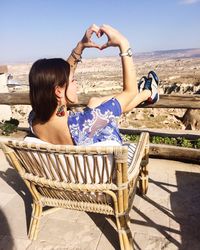 Rear view of woman gesturing heart shape while sitting on chair against sky