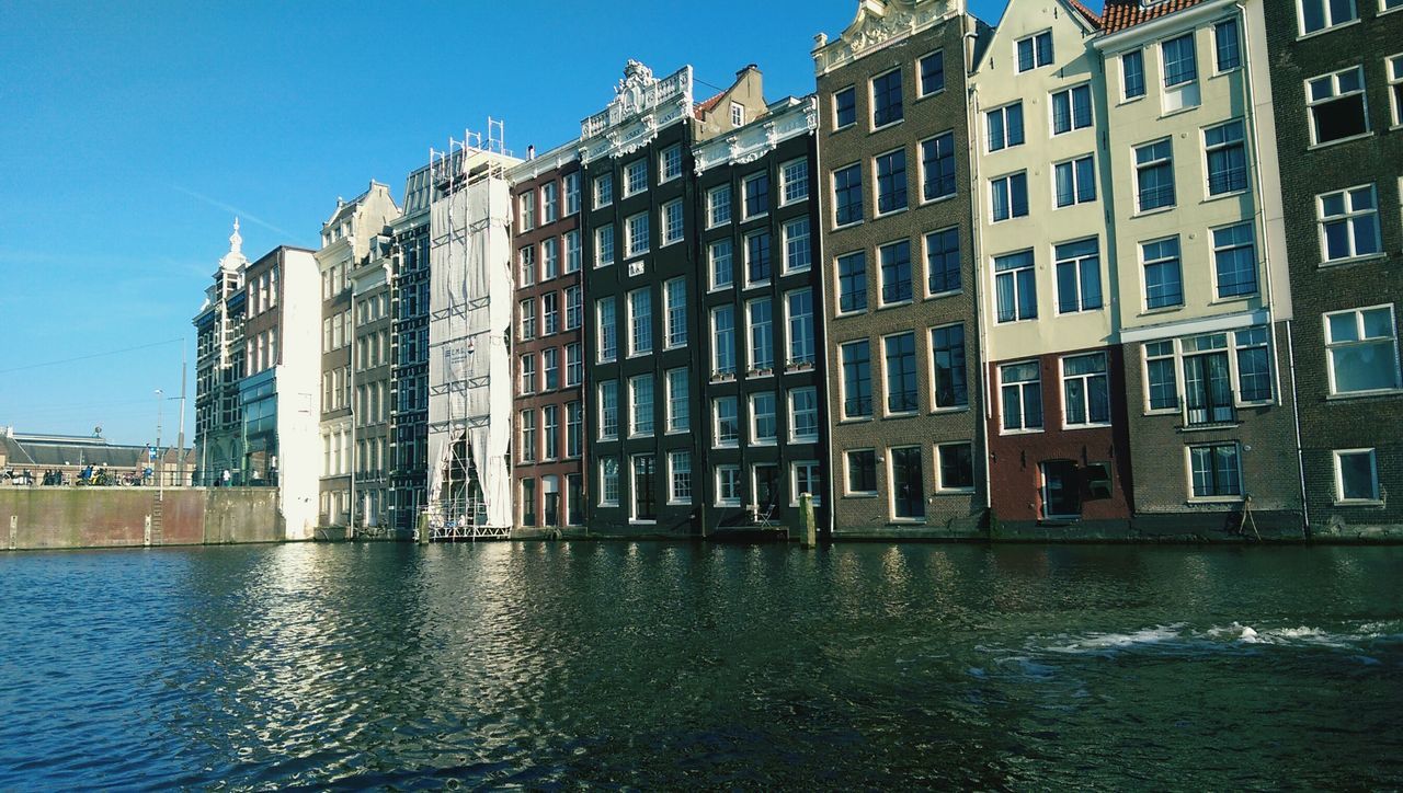 architecture, building exterior, built structure, water, waterfront, reflection, building, blue, canal, residential building, city, window, residential structure, rippled, clear sky, sky, day, sunlight, outdoors, river