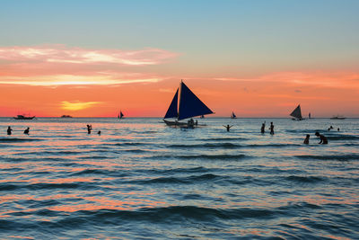 Silhouette people with sailboat in sea against orange sky