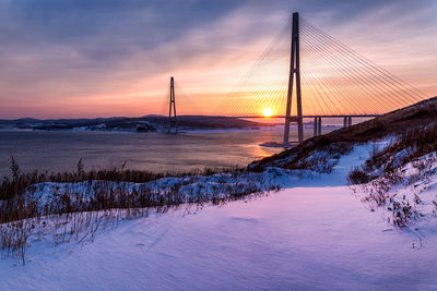 Bridge over snow covered land against sky during sunset