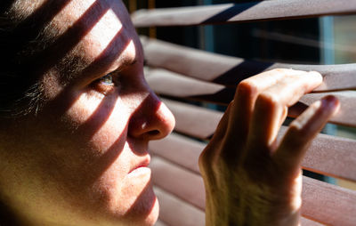 Adult woman with blue eyes opening a wooden curtain with her fingers to look through the window while the sunlight creates sun and shadow on her face. horizontal photo.