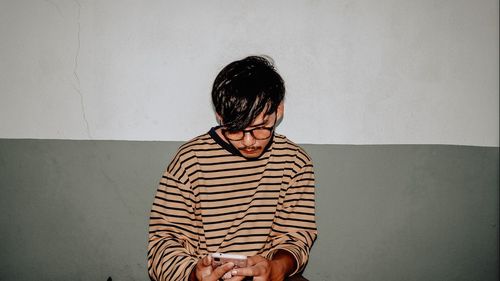 Young man using phone while sitting against wall at home